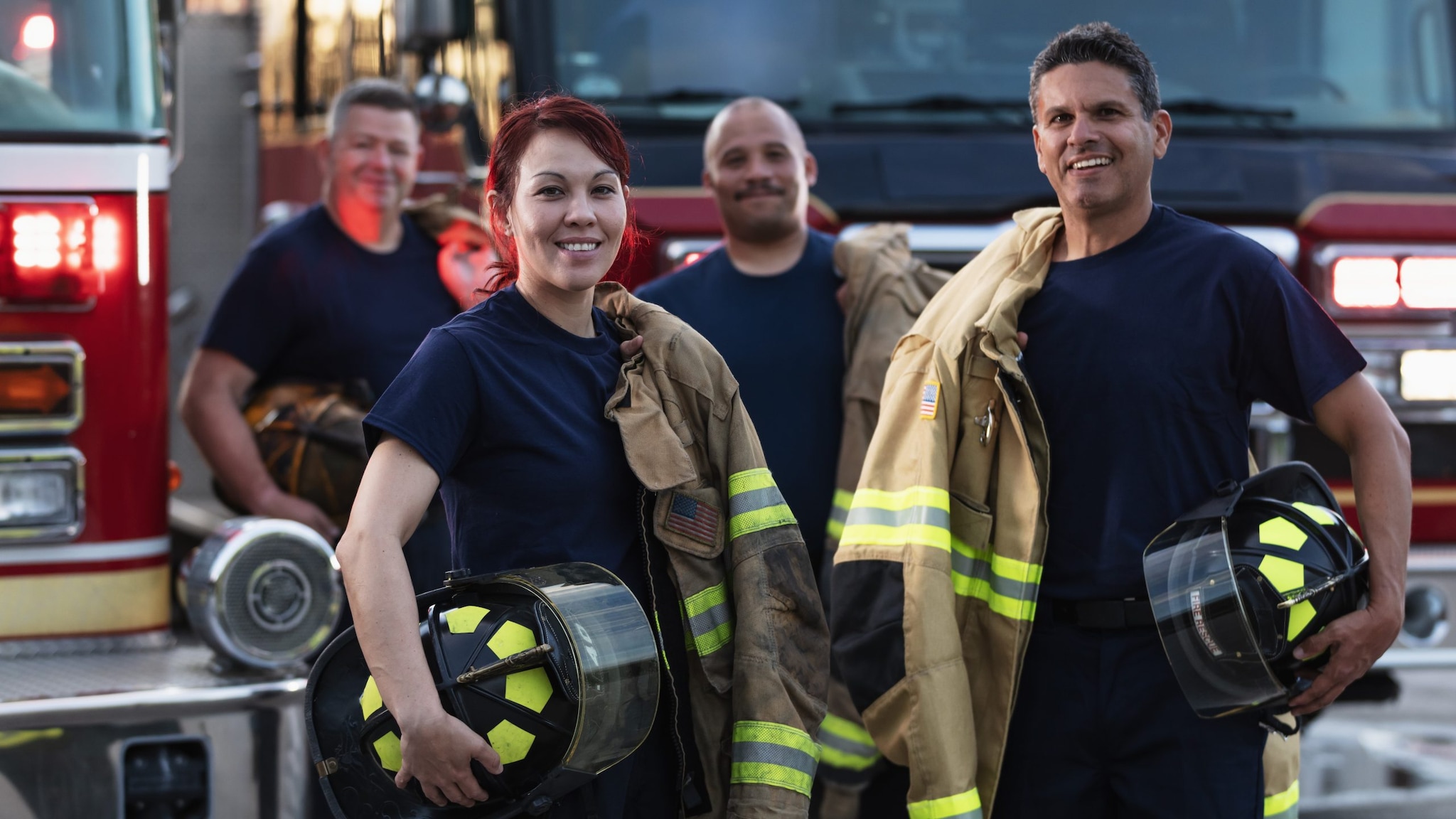 A multiracial group of four firefighters standing in front of fire engines holding PPE