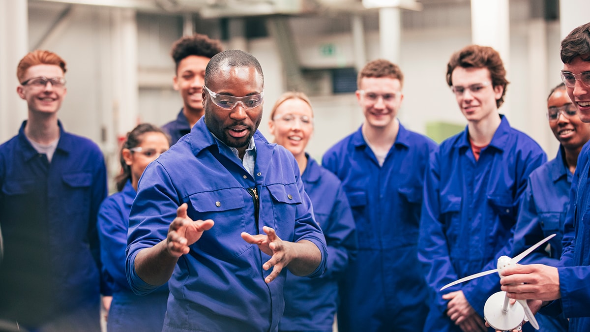 Group of people wearing coveralls watching someone give a demonstration.