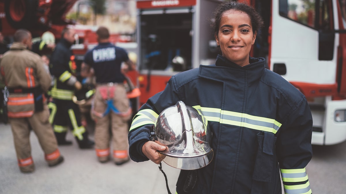 Bombera embarazada en primer plano, con otros bomberos al fondo.