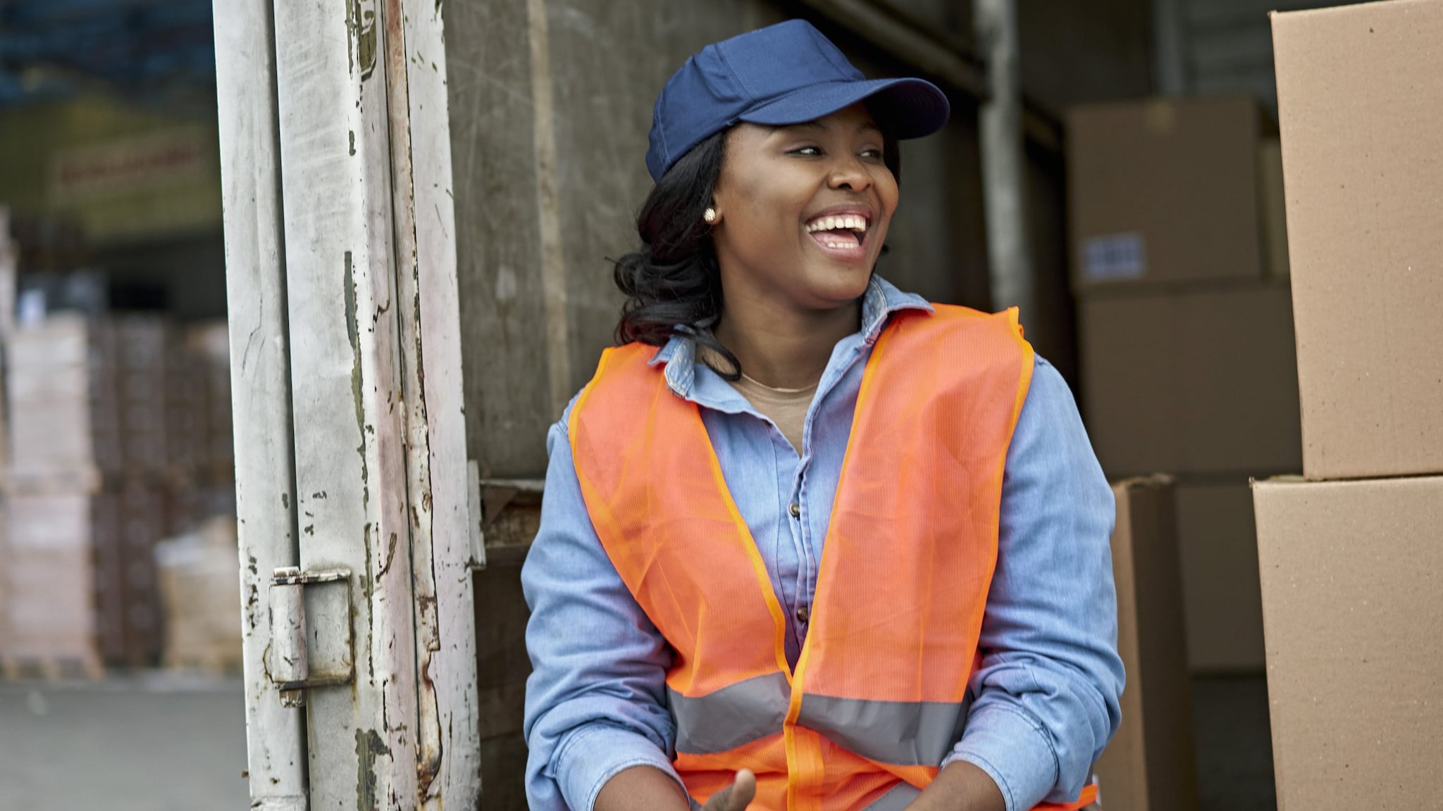 woman wearing blue baseball hat and orange safety vest sitting on the back of a truck with boxes