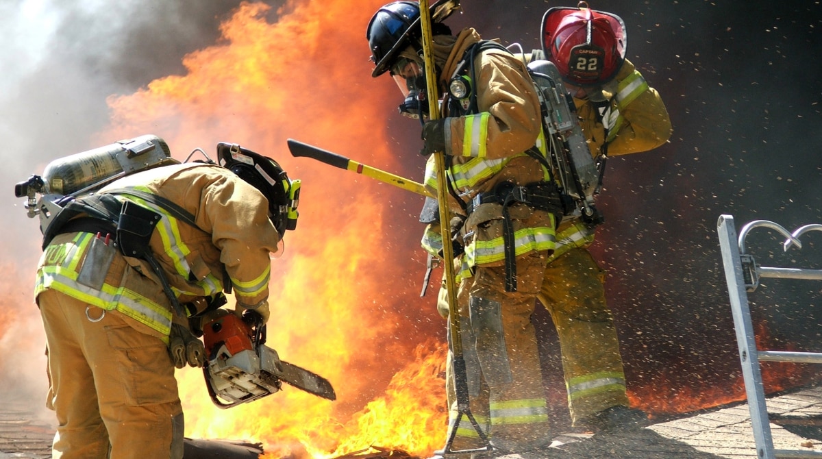 Firefighters wearing PPE, carrying gear, near large flames.