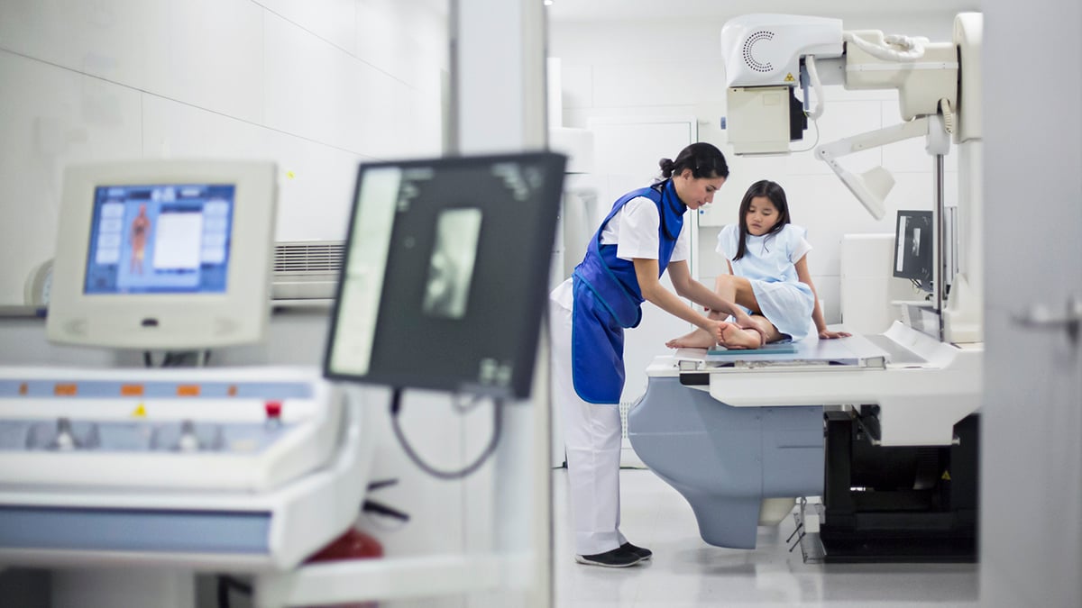 A radiology technologist wearing protective lead drapes positions a child for an x-ray