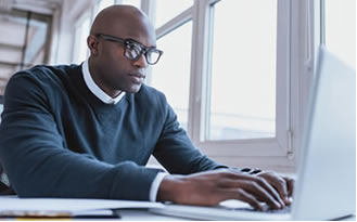 Man wearing glasses and a sweater, working on a laptop at a desk near a window.