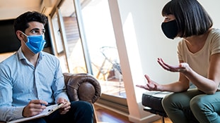 A seated, masked male behavioral health professional talking with a seated, masked female patient.