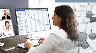 Woman in white shirt sitting at desk looks at a video conference call on one monitor and a spreadsheet on another monitor.