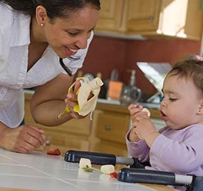 A mother interacting with her 6 month old daughter as she feeds her a banana.