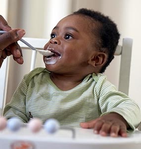 6 month old baby sitting in high chair and being fed solid foods for the first time from a spoon.