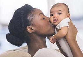 A mother raising her infant towards her and kissing her. The infant has soft, tranquil face.