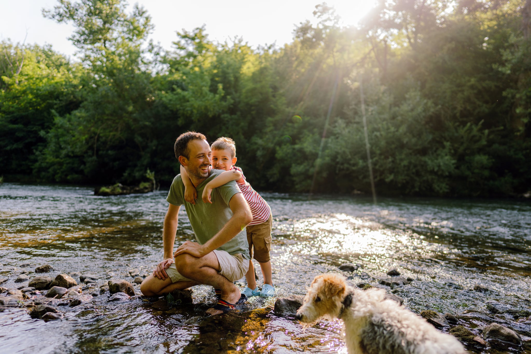 A photo of a man kneeling down while a boy hangs on his neck. Both are smiling and are standing on a shallow part of a stream water. A dog stands close by. The sun shines through lush green trees in the background.