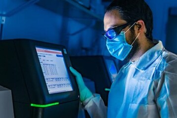 A scientist wearing biosafety level 2 PPE gear including gloves, laboratory coat, goggles, and mask, using a sequencing instrument.