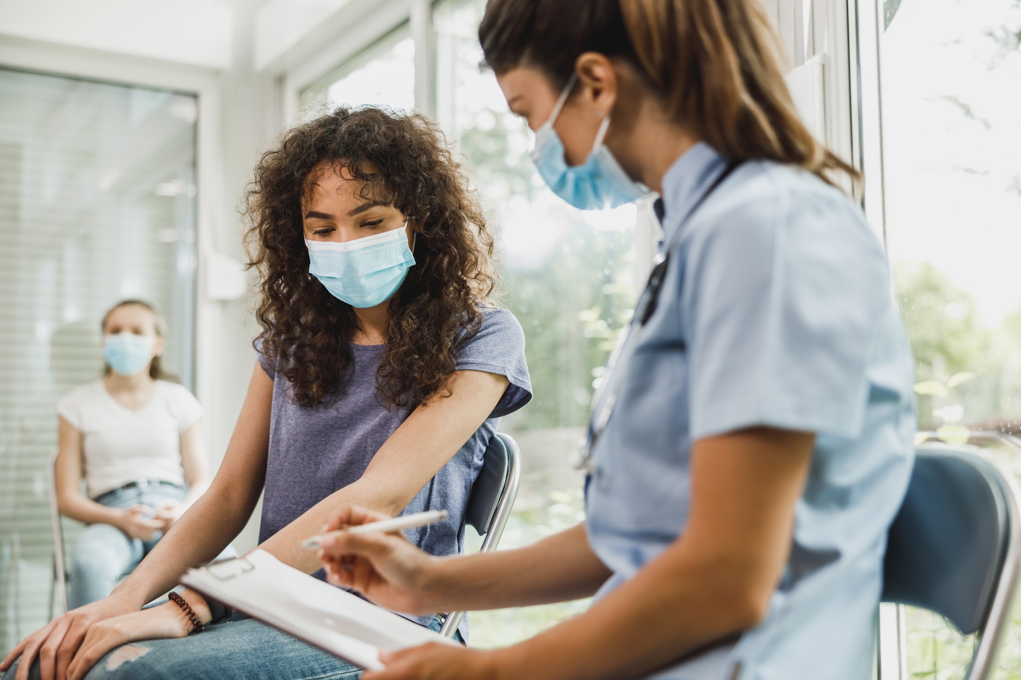 A clinician writes on a notepad while meeting with a patient in an open clinic setting.