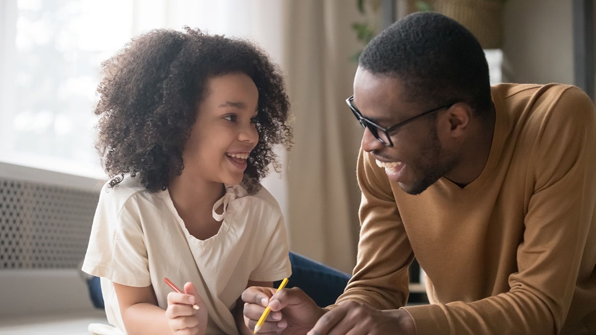 A father sits beside his young daughter to help her color a picture.