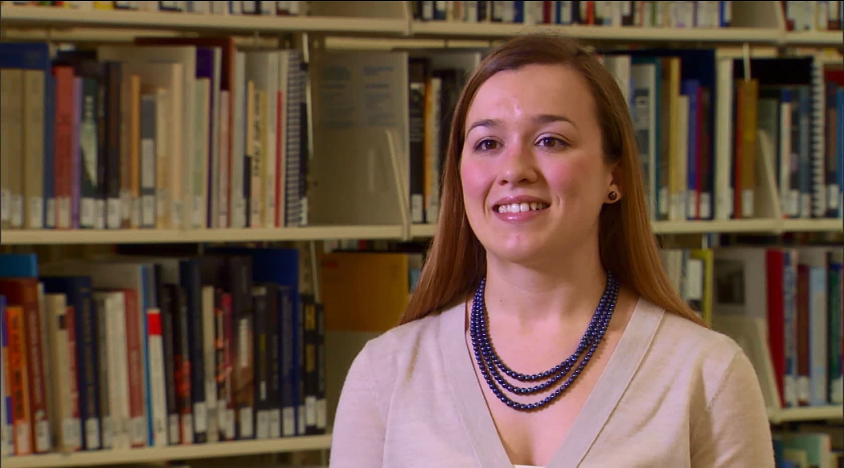 A pleasantly smiling woman wearing a purple necklace and earrings surrounded by books in a library.