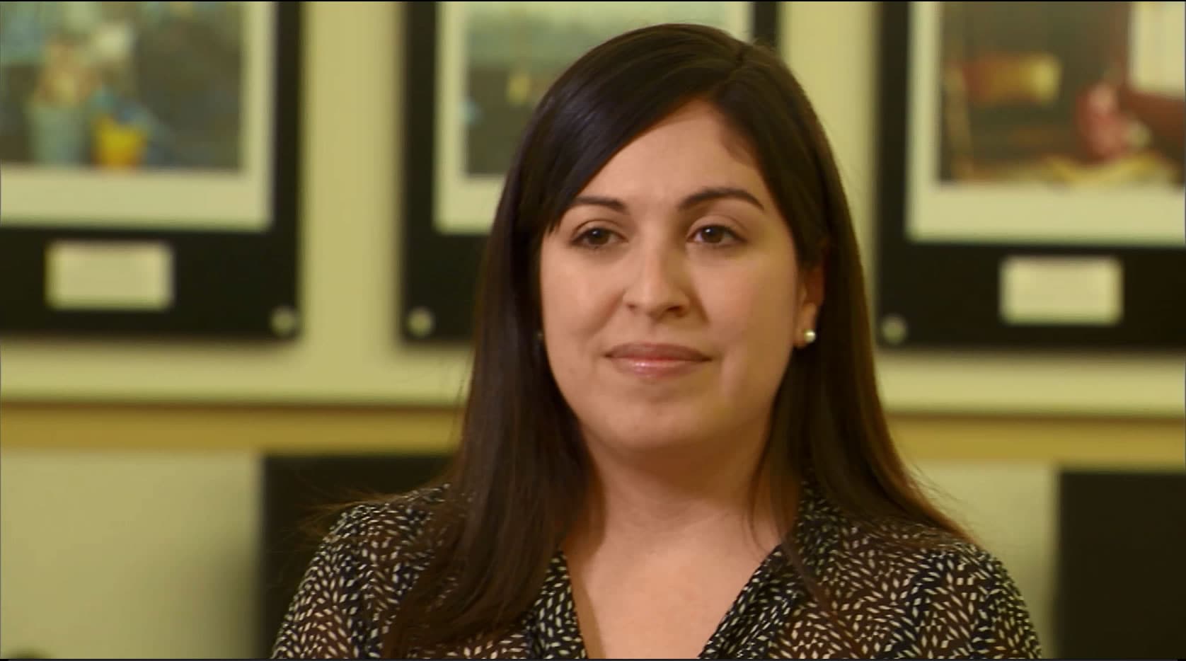 A dark-haired woman in a polka dot button down shirt in an office setting with computers and pictures hanging on the wall in the background.