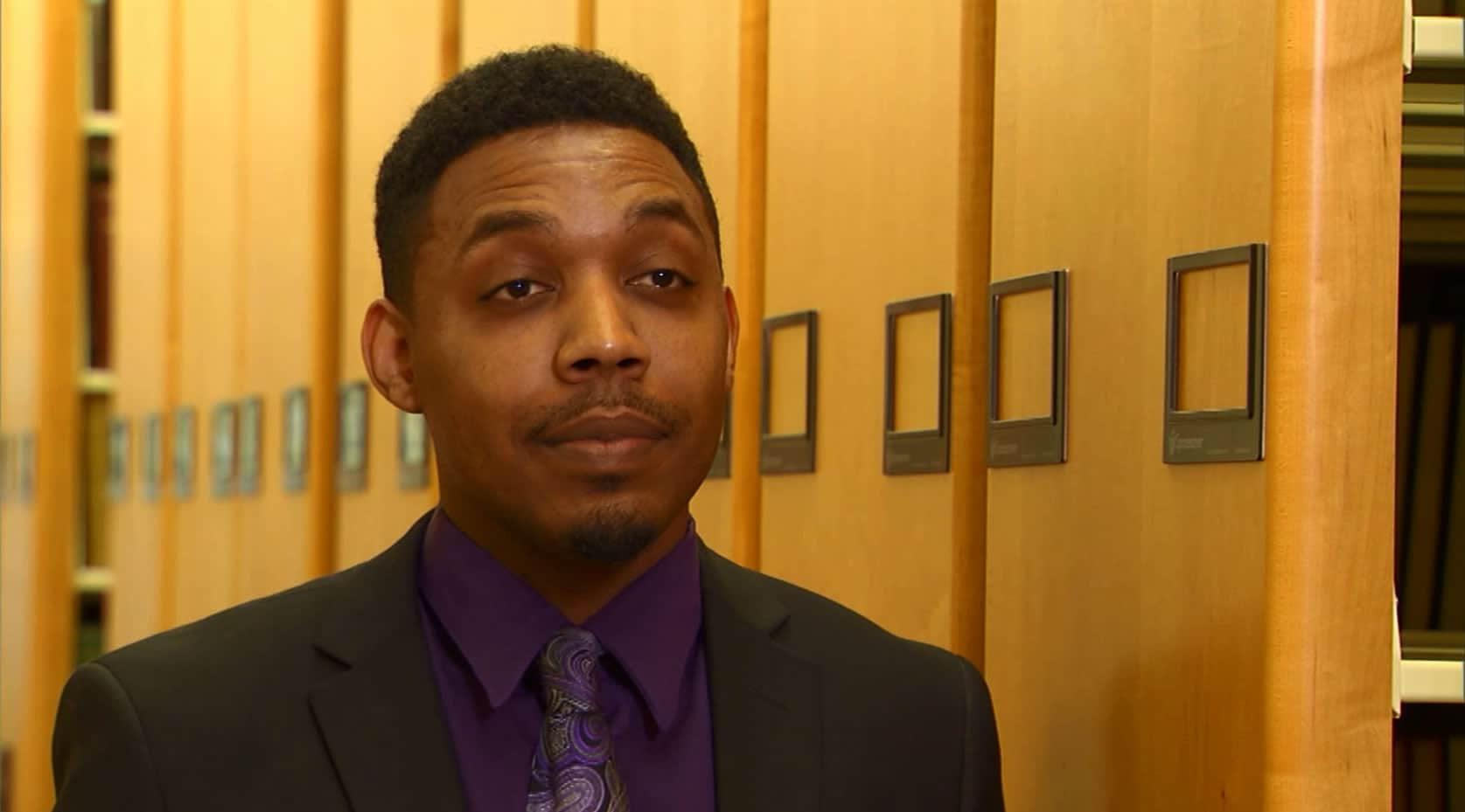 A man in a suit and tie standing in front of a row of bookshelves, looking at the camera with a confident smile.