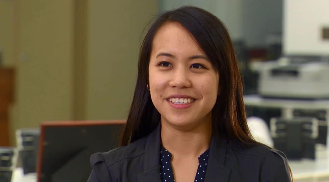 A woman smiling with long dark hair, wearing a polka dot top and black blazer, sitting in front of a computer screen.