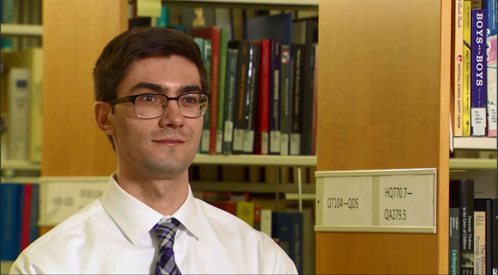 A dark-haired man, wearing glasses in a white shirt and tie, sits in a library setting surrounded by books on shelves.