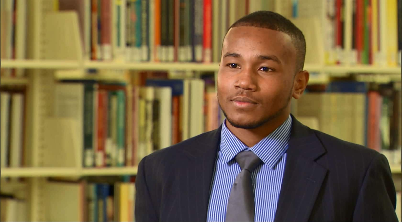 A young man in a suit and tie standing in front of bookshelves, looking professional and sophisticated.