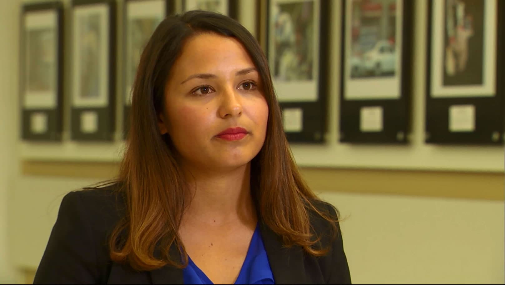 A woman wearing a blue shirt and black blazer with a wall of hanging pictures in the background.