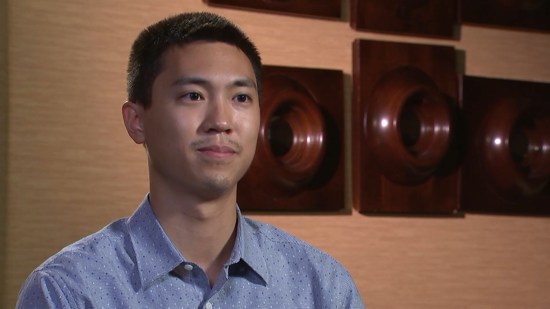 A young man in a blue shirt sitting in front of a wall.