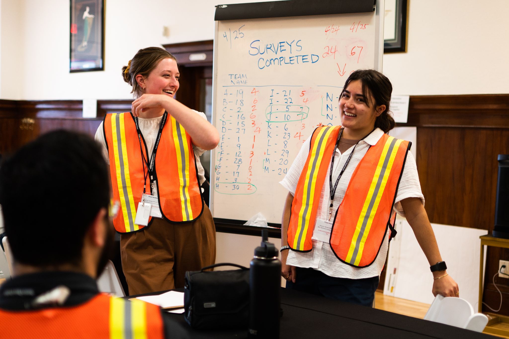 Two Public Health Associates wearing high-visibility reflective vests smile while presenting survey data to colleagues.