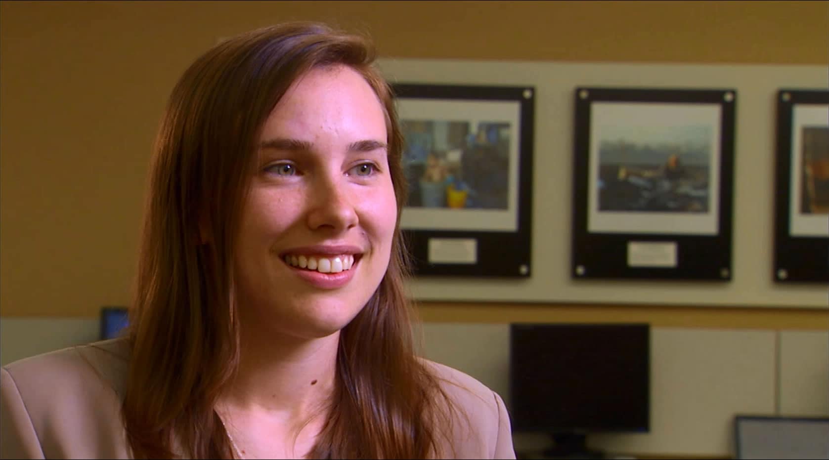 A woman looking happy and engaged in an office setting in front of a computer screen with pictures hanging on the wall.