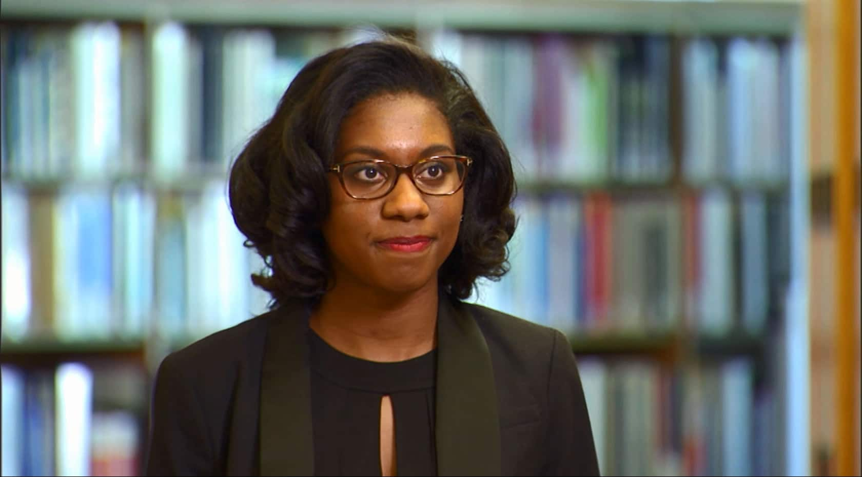 A woman wearing glasses with short dark hair, in a dark colored blazer stands in front of blurred bookshelves full of books in the background.