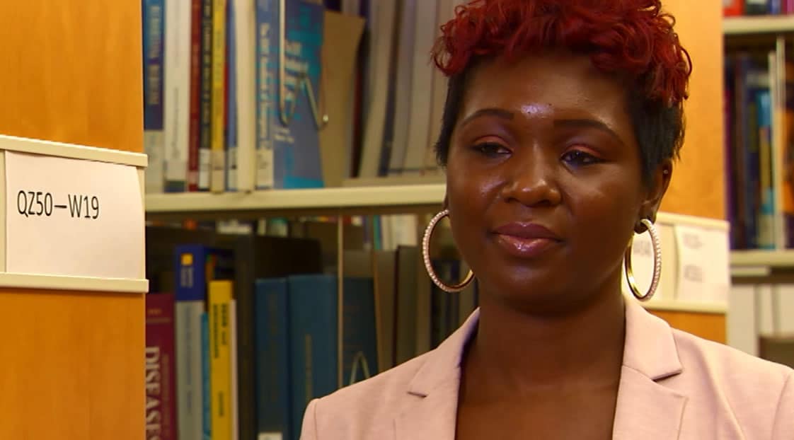 A woman with red hair, wearing large gold hoop earrings and a light pink blazer, sitting in a library, surrounded by books.