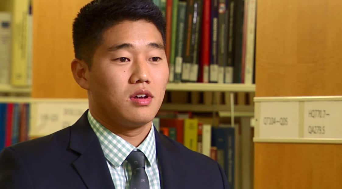 A professional young man in a suit and tie standing in front of bookshelves in a library setting.