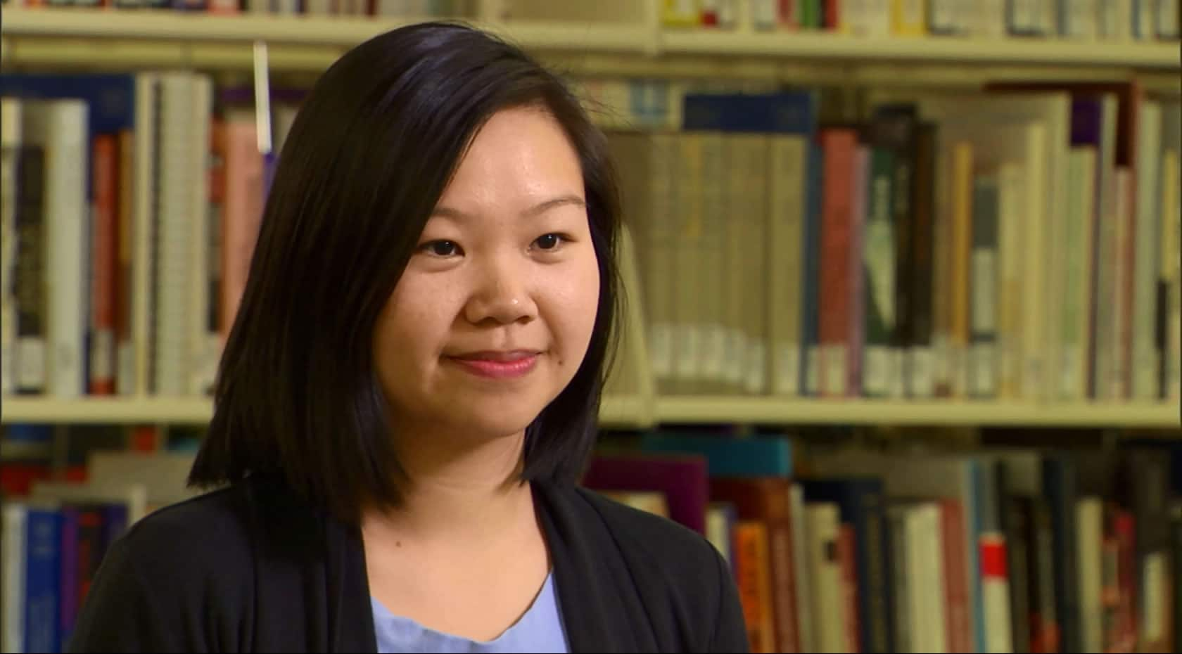 A young woman in business clothing in from of a shelf of books looking confidently at the camera.