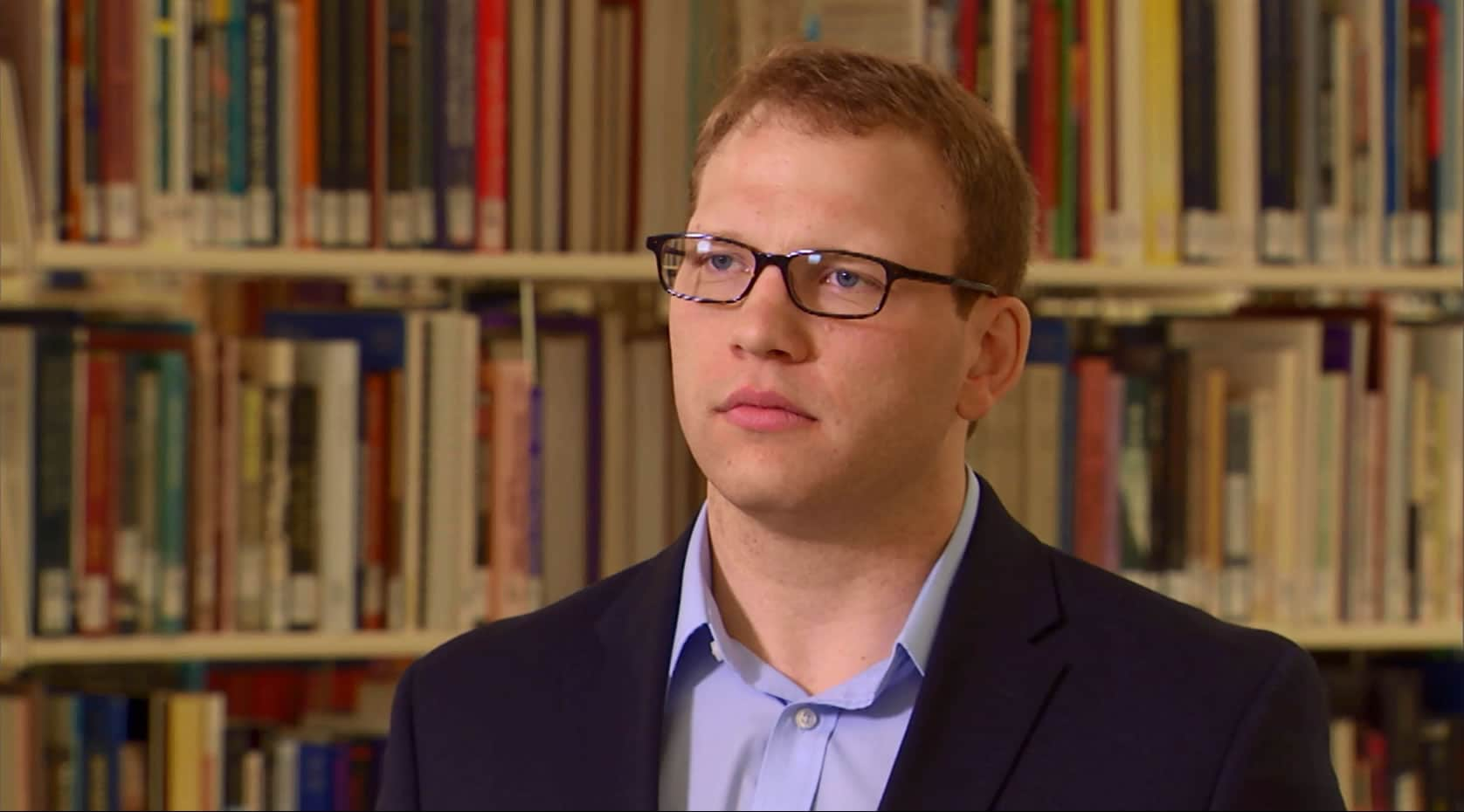 A bespectacled man standing in of a library, surrounded by shelves of books.