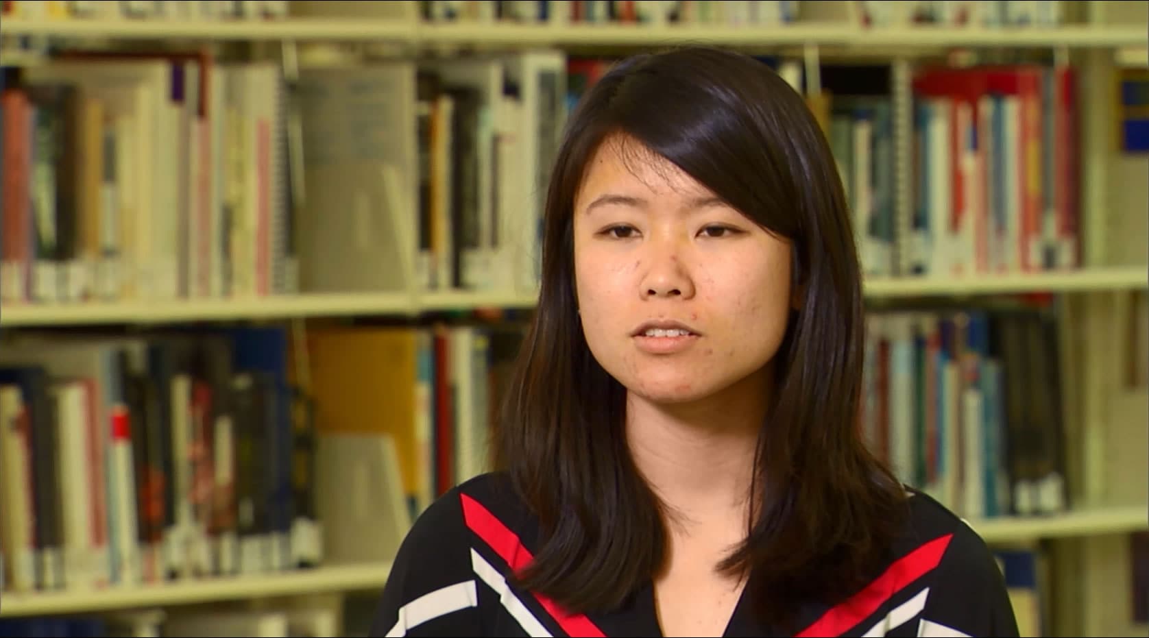 A young woman wearing a red and white striped shirt in a library, surrounded by bookshelves filled with books.