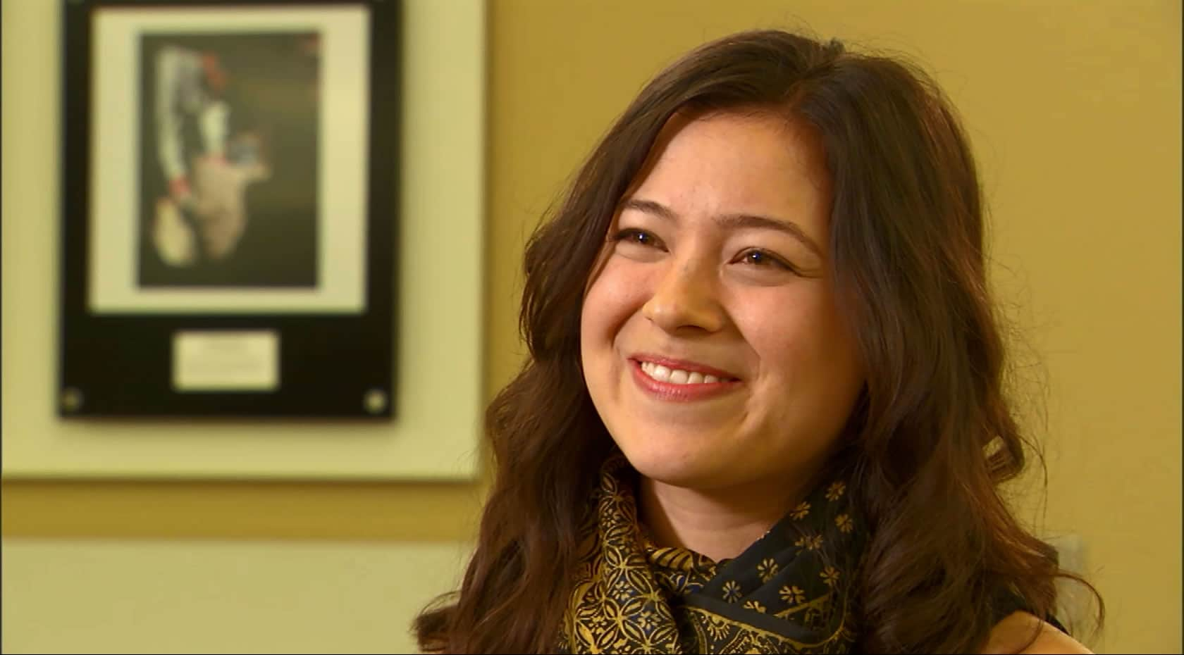 A woman wearing a scarf and black shirt smiles warmly, radiating happiness and contentment in front a hanging picture on a mustard-colored wall.