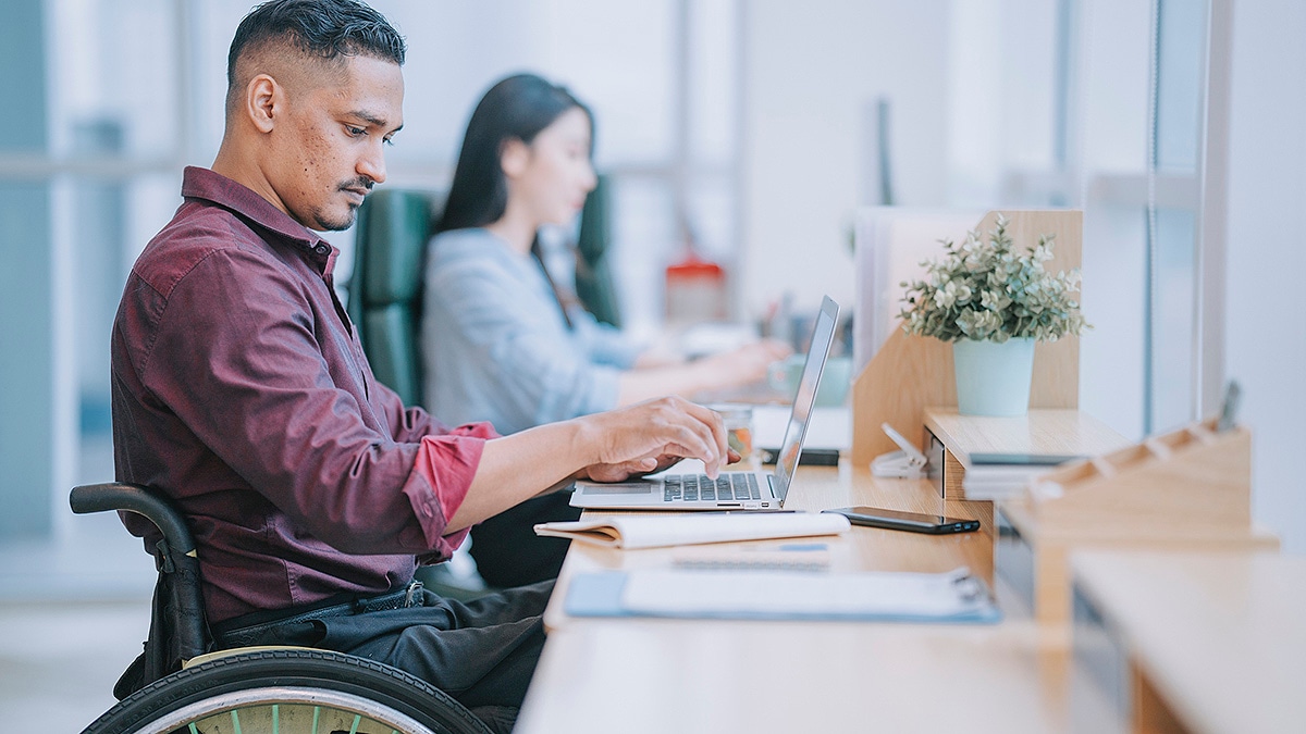 Man in a wheelchair sitting at a long table working on a laptop. Woman in background also working.