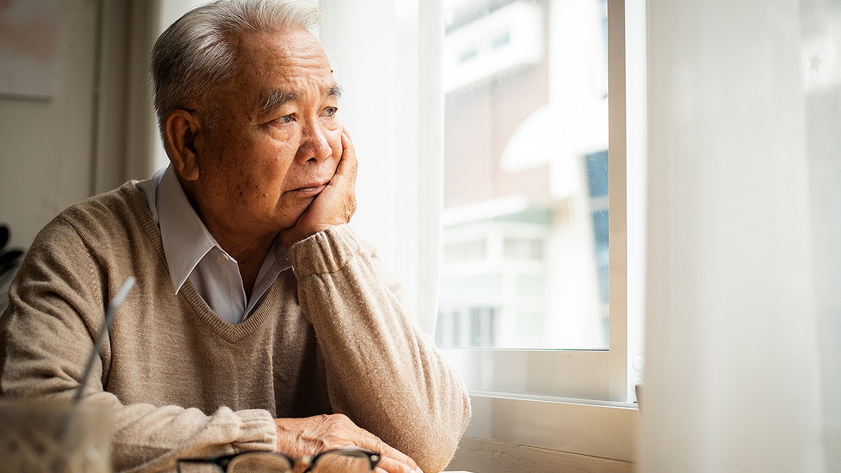 Older, isolated, Asian man sadly looking out window with head in hand.