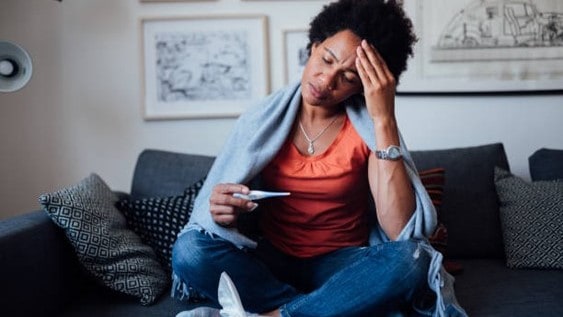 Woman sitting on the couch holding a thermometer with a hand to her forehead