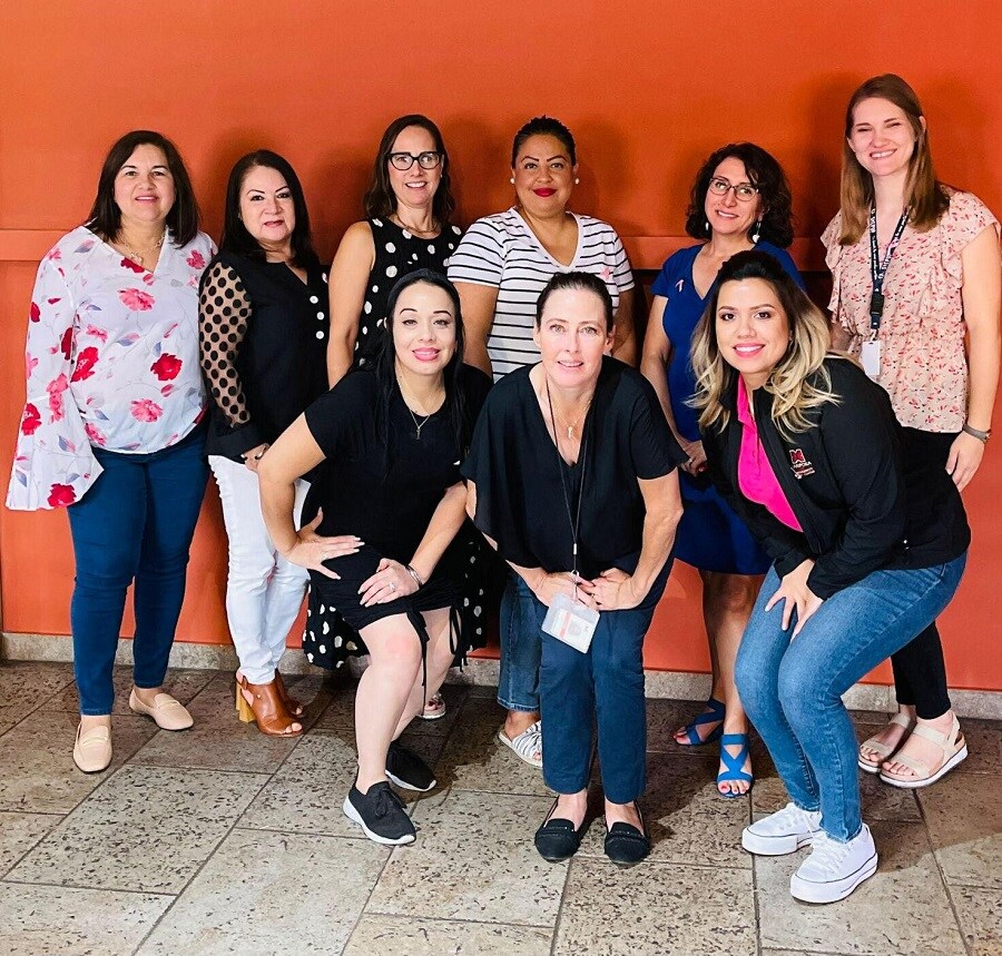 Group of women standing in front of a wall. There are three women in front and six women in the back. The women are of varying ages.