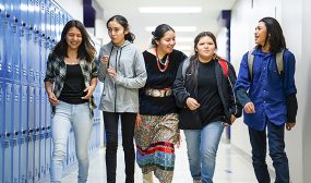 five high schoolers walking down a corridor at school while talking to each other.