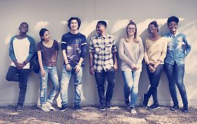 Group of diverse friends standing against a white wall outside while sun shines down on them