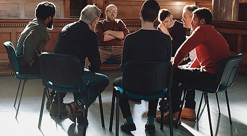 Group of sad, young, male adults in a darker room, having a group therapy session.