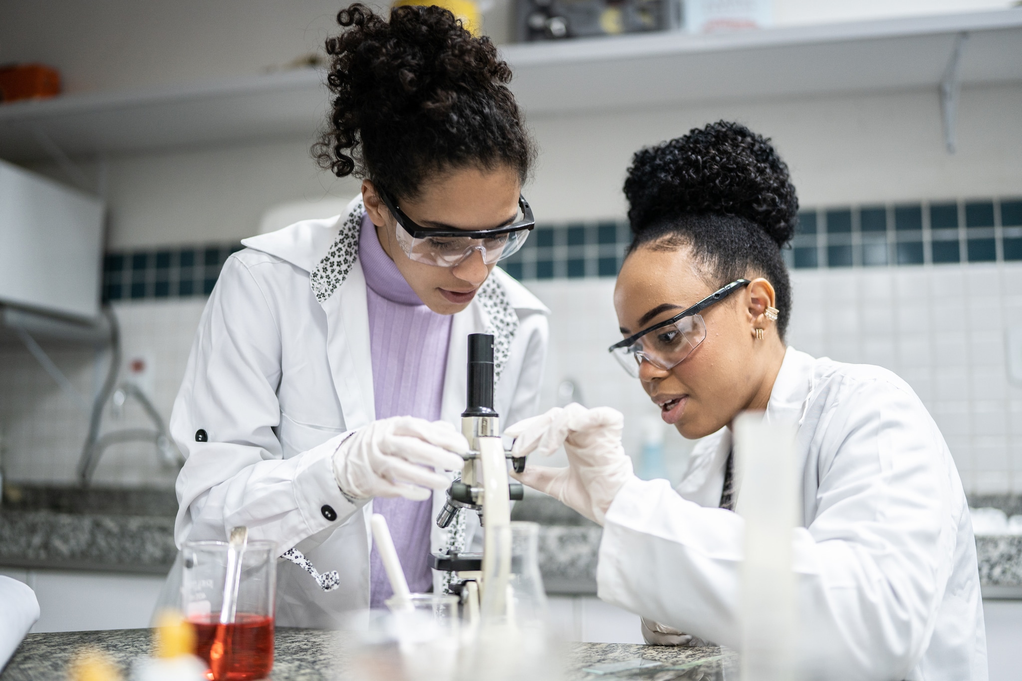 Two people wearing white coats and goggles look at a microscope in a lab setting