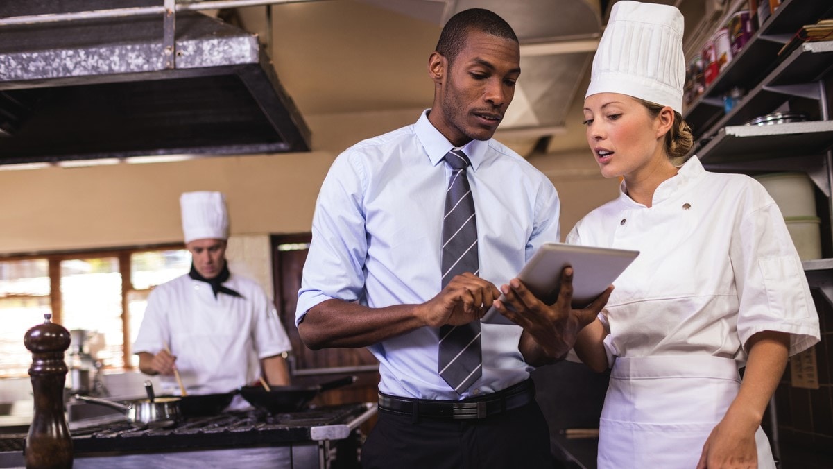 Investigator holding a clipboard talks to a chef in a restaurant kitchen.