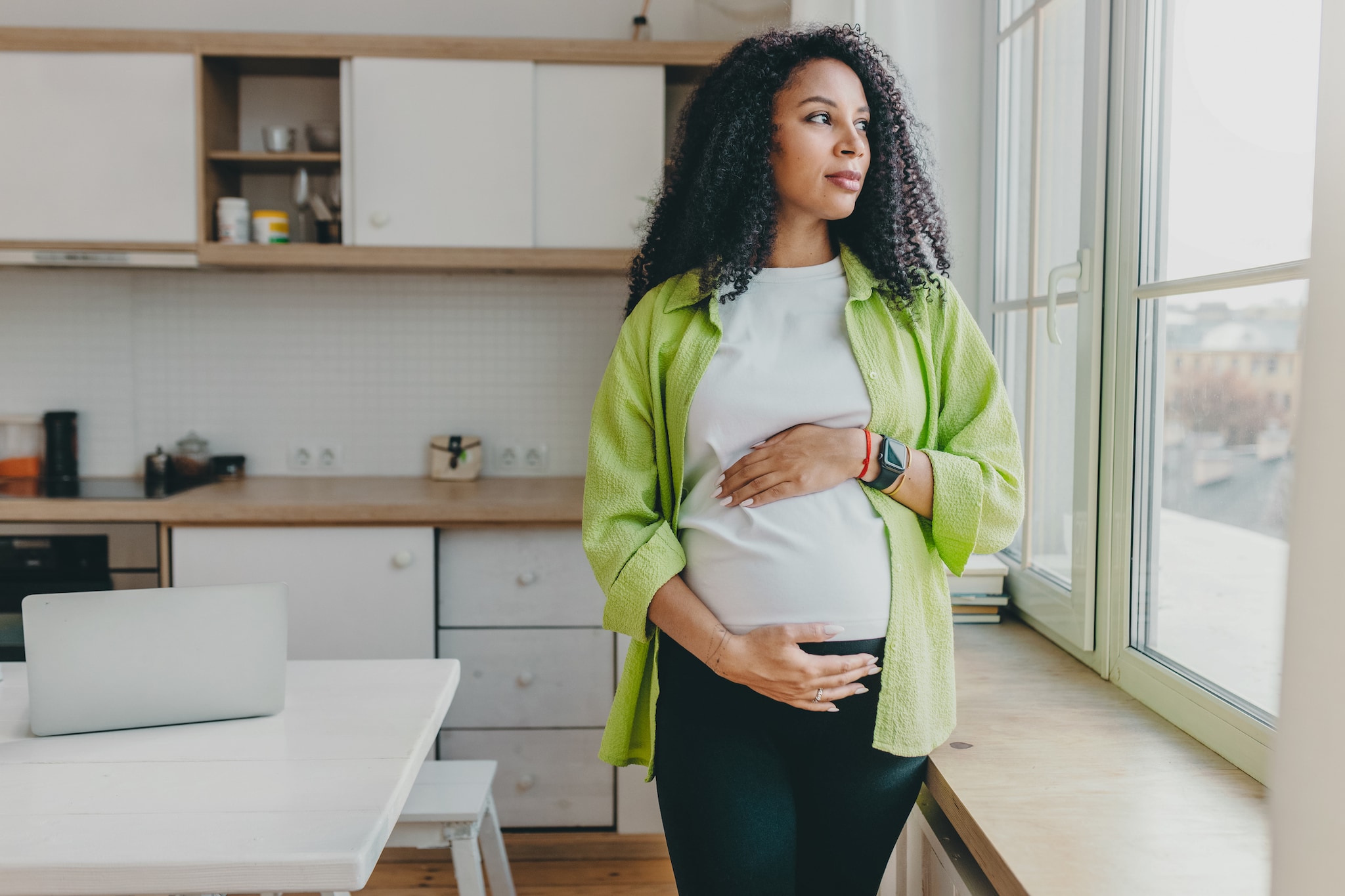 Pregnant person of color standing and looking out the kitchen window, while holdingbelly.