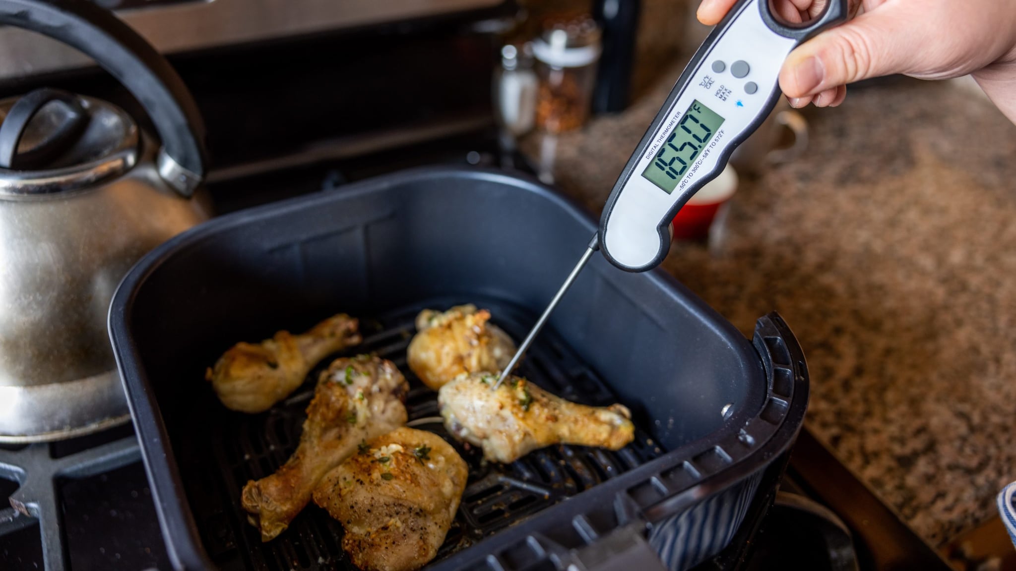 Person inserting food thermometer into chicken pieces in an air fryer to check temperature. Thermometer reads 165°F.