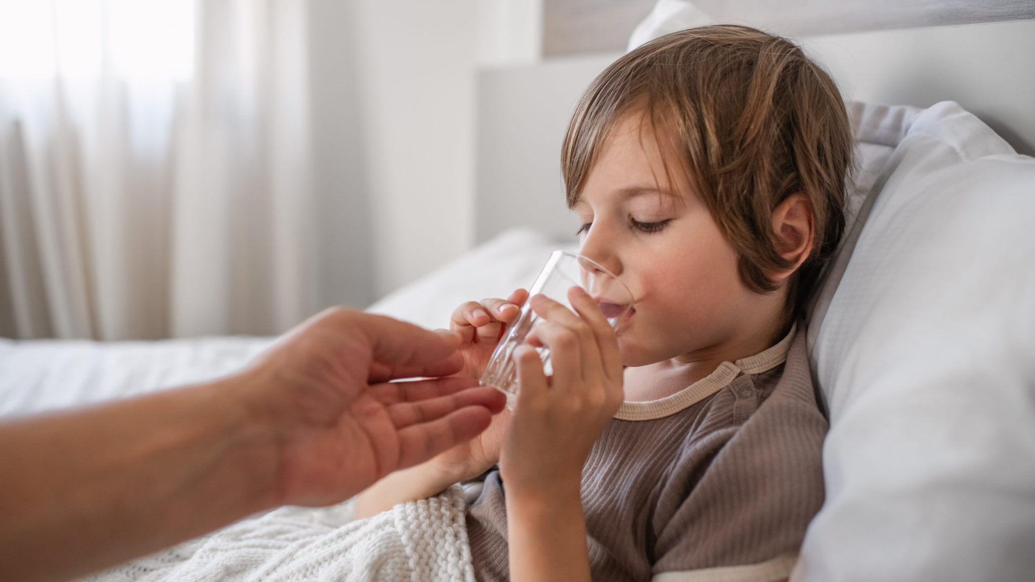 A child propped up in bed drinking water. The glass is being supported by a man's hand.