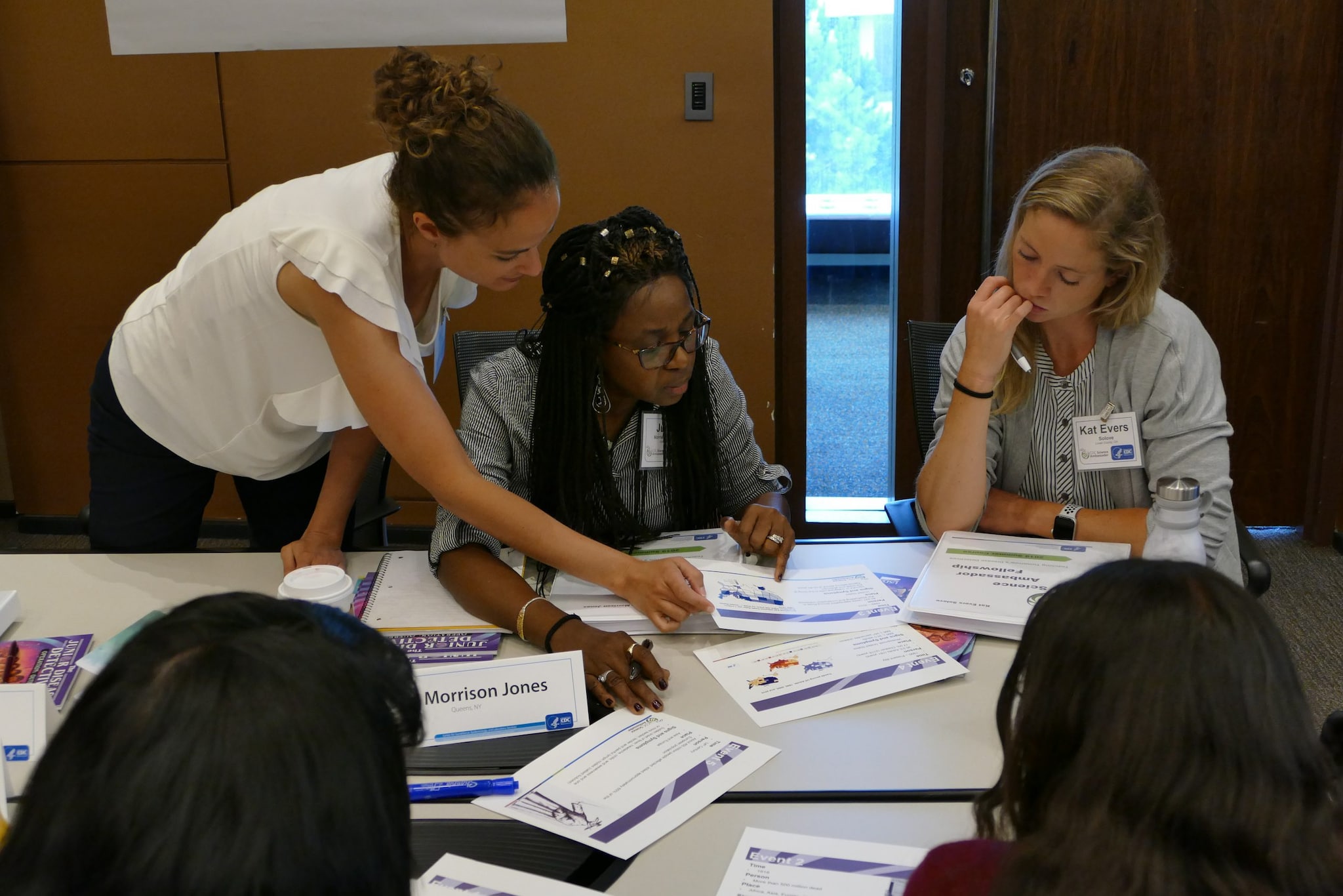 A dark-haired woman in white shirt works with other teachers. They are at a table covered in educational materials.