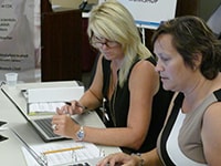 A blonde-haired woman in a black tank top sits at a table in front of a laptop looking at a dark-haired woman's laptop next to her.