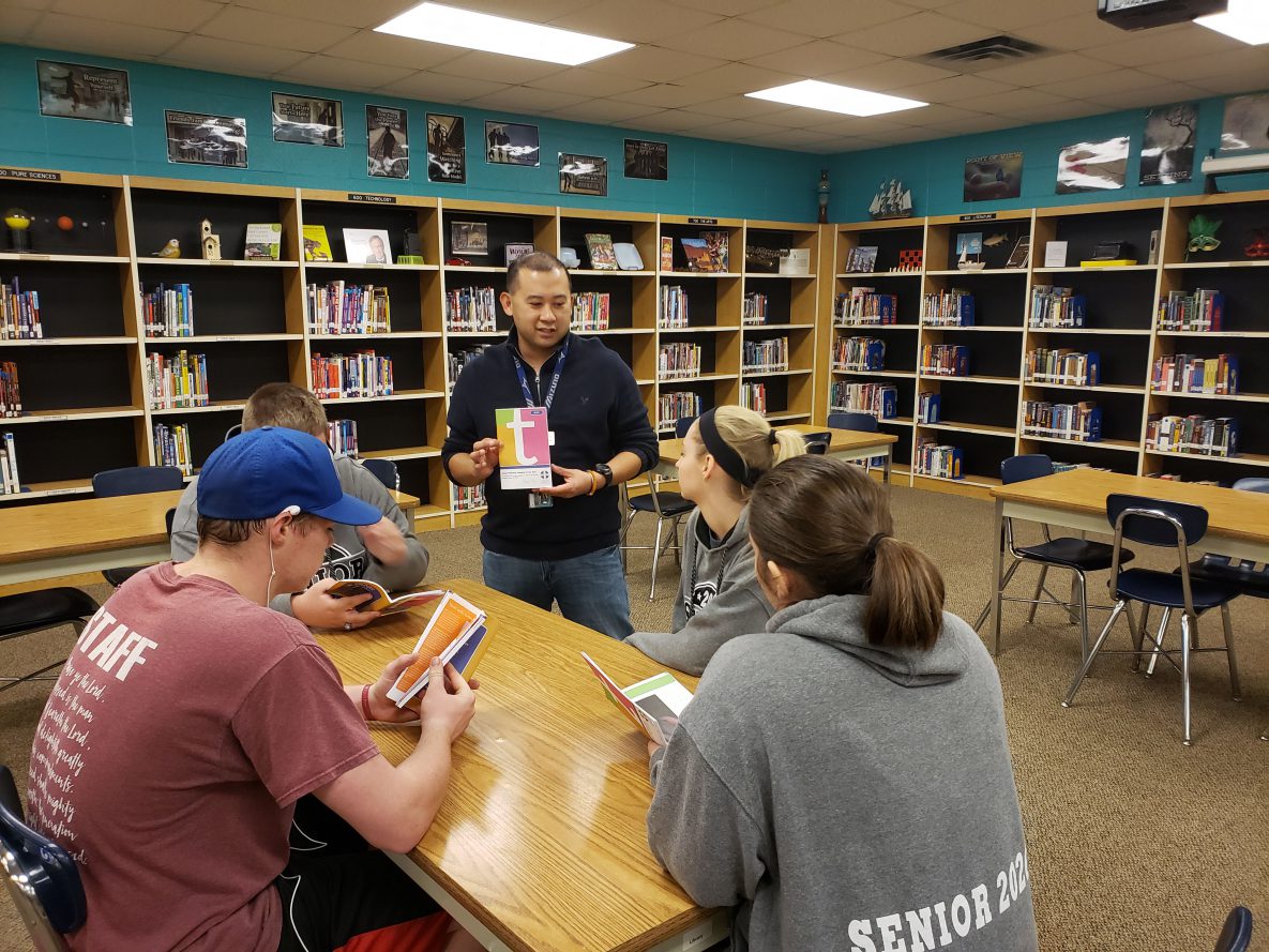 Man standing in a library showing a book to others.