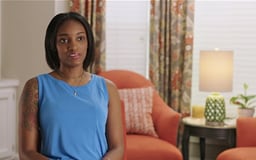 Black Female sitting in room with window and chair behind her talking to camera