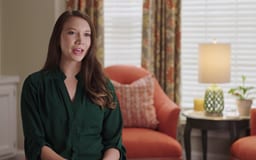 White female sitting in room with window and chair behind her talking to camera